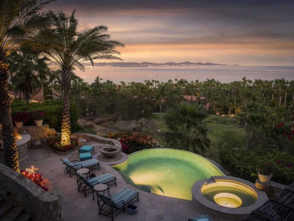 A glowing hillside pool sits under tall palm trees with a soft sunset over the ocean. Warm lights highlight the terraces and gardens, creating a dreamy, peaceful mood. The ocean views stretch all the way to the horizon, making the whole scene feel luxurious and calm.