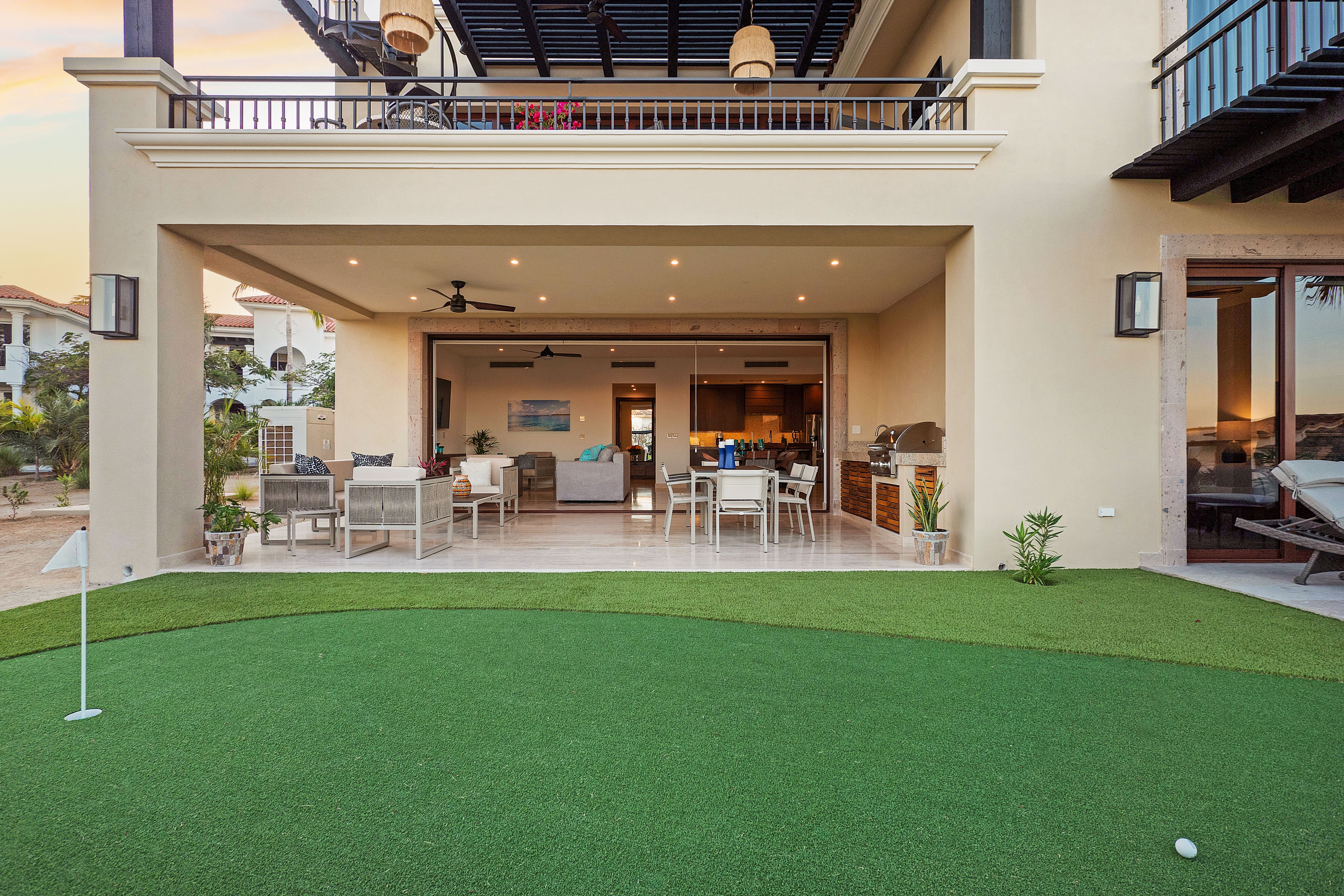 Covered patio with outdoor lounge seating, dining area, built-in grill, and direct access to a private putting green, framed by a two-story luxury villa.
