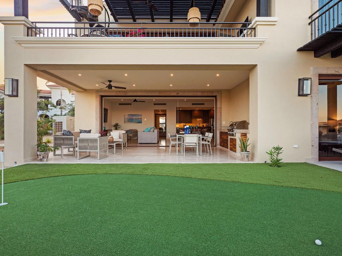 Covered patio with outdoor lounge seating, dining area, built-in grill, and direct access to a private putting green, framed by a two-story luxury villa.
