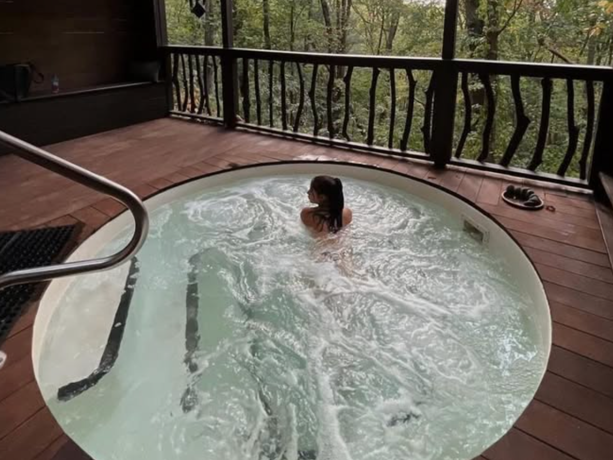 A woman relaxes in a round outdoor hot tub on a wooden deck surrounded by trees and forest views.