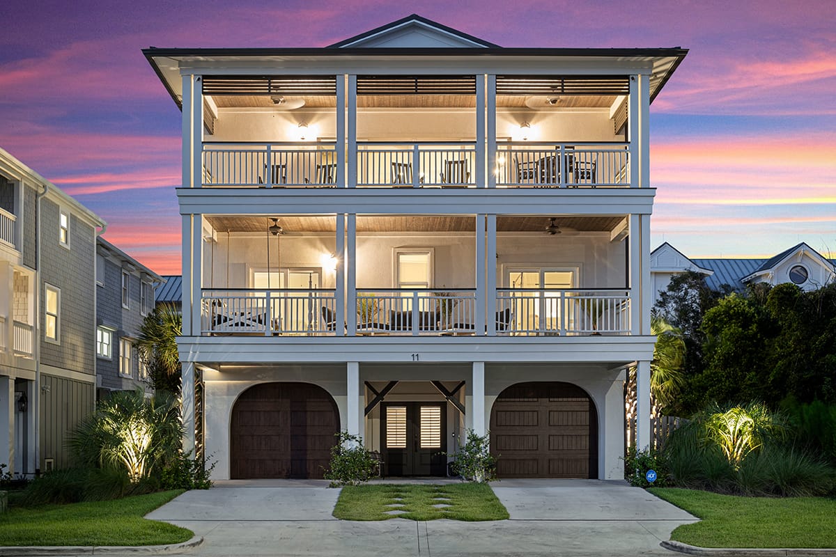 A modern three-story beach house with glowing lights and spacious balconies on each level, framed by palm trees and set against a vibrant purple and pink sunset sky.