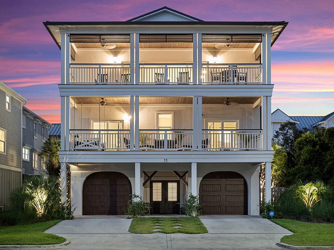 A modern three-story beach house with glowing lights and spacious balconies on each level, framed by palm trees and set against a vibrant purple and pink sunset sky.