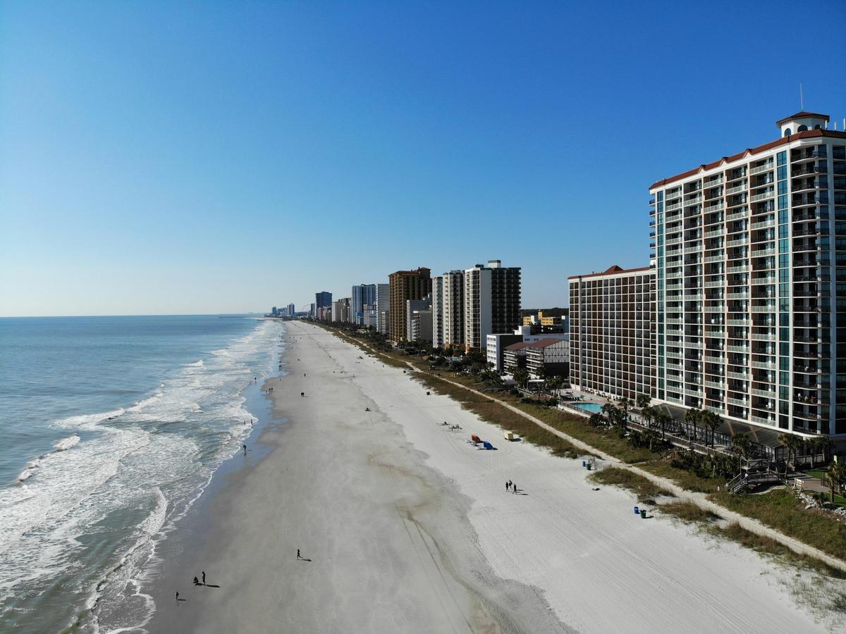 Aerial view of a long sandy beach lined with tall oceanfront hotels and resorts under a bright blue sky, with gentle waves rolling onto the shore.