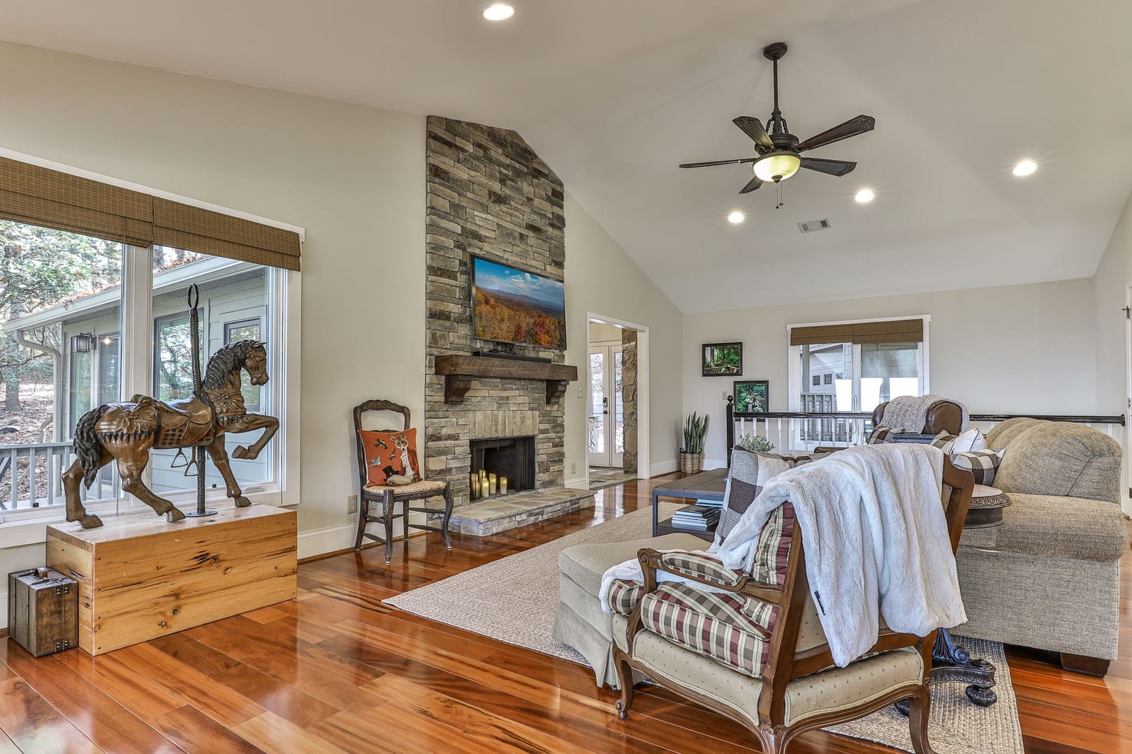 A bright, inviting living room with hardwood floors, a stone fireplace with mounted TV, neutral-toned seating, vaulted ceilings with a ceiling fan, and rustic accents overlooking a wooded outdoor area.