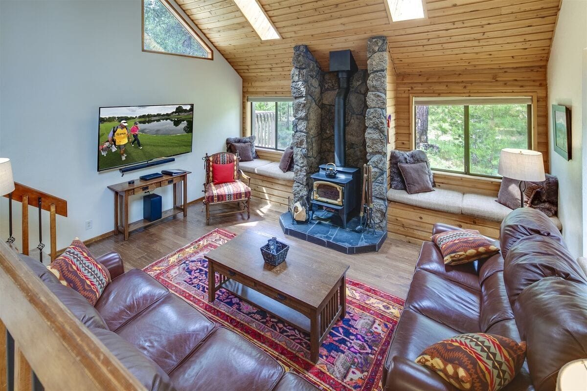 A cozy cabin-style living room with warm wood finishes, leather sofas, and a stone fireplace centerpiece perfect for chilly nights. Large windows and skylights fill the space with natural light, while plush seating invites guests to unwind after a day outdoors. The blend of rustic charm and comfort makes it ideal for family gatherings.
