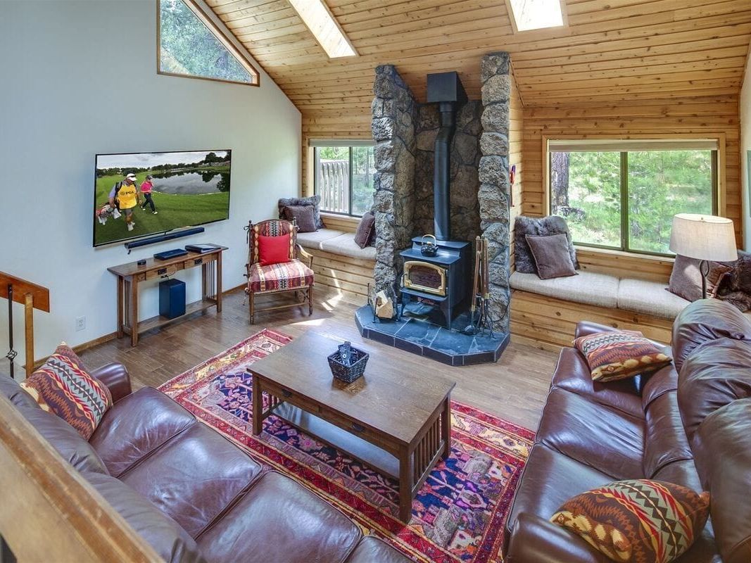 A cozy cabin-style living room with warm wood finishes, leather sofas, and a stone fireplace centerpiece perfect for chilly nights. Large windows and skylights fill the space with natural light, while plush seating invites guests to unwind after a day outdoors. The blend of rustic charm and comfort makes it ideal for family gatherings.