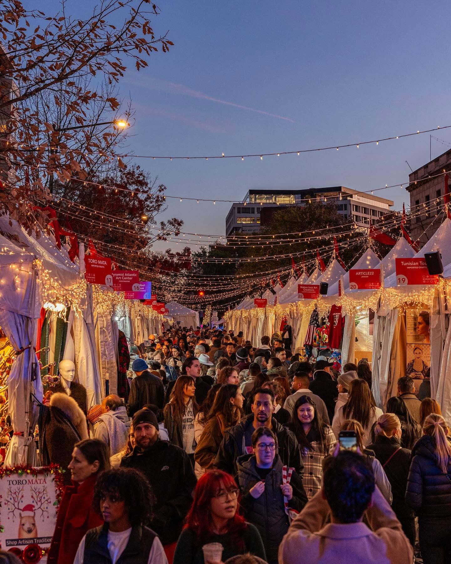 A dense crowd of holiday shoppers fills the string-light-draped aisles of the Downtown Holiday Market in Washington DC at dusk, passing rows of white vendor tents selling imported crafts and artisan goods. 