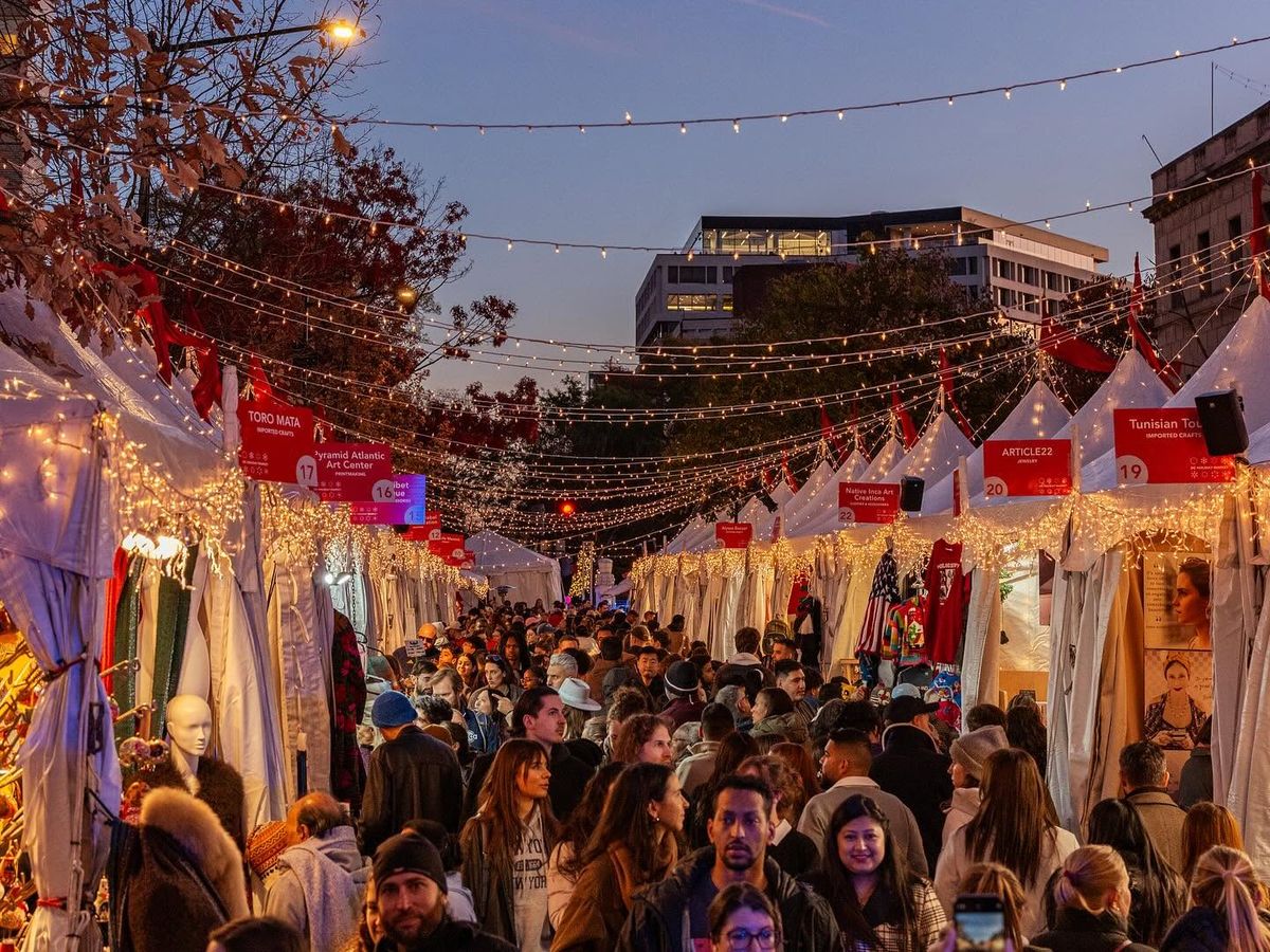 A dense crowd of holiday shoppers fills the string-light-draped aisles of the Downtown Holiday Market in Washington DC at dusk, passing rows of white vendor tents selling imported crafts and artisan goods.