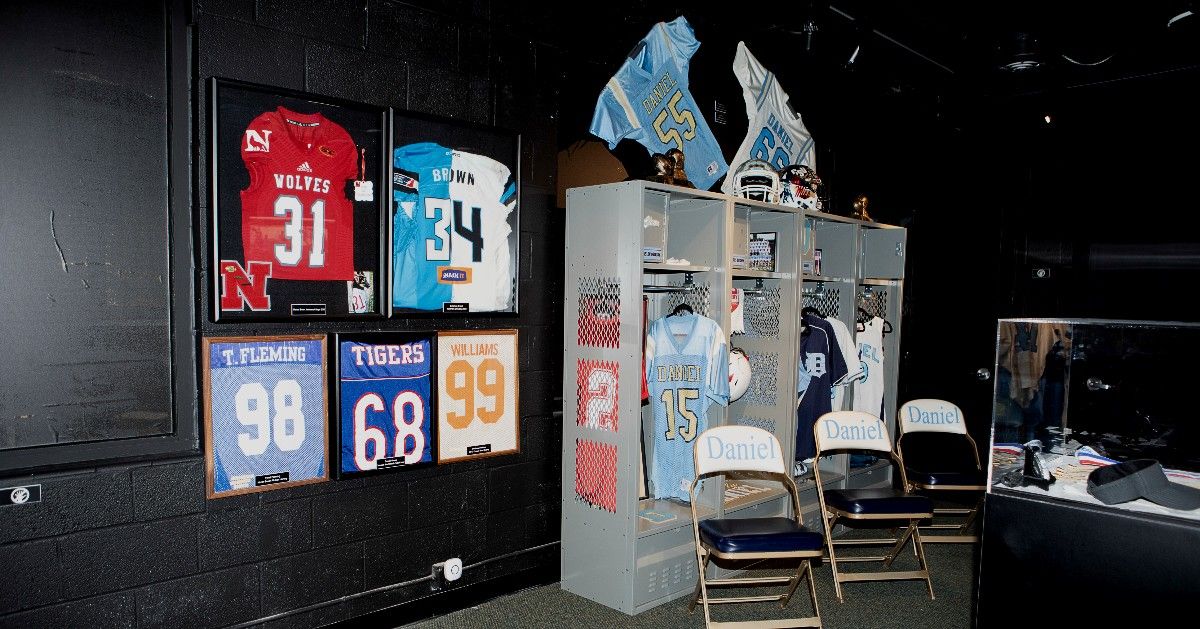 A sports memorabilia exhibit at the Clemson Area African American Museum displays framed football jerseys from local athletes mounted on dark walls alongside a recreated locker bay with Daniel High School uniforms, helmets, and folding chairs. The installation honors the athletic achievements of African American players from the Clemson area community