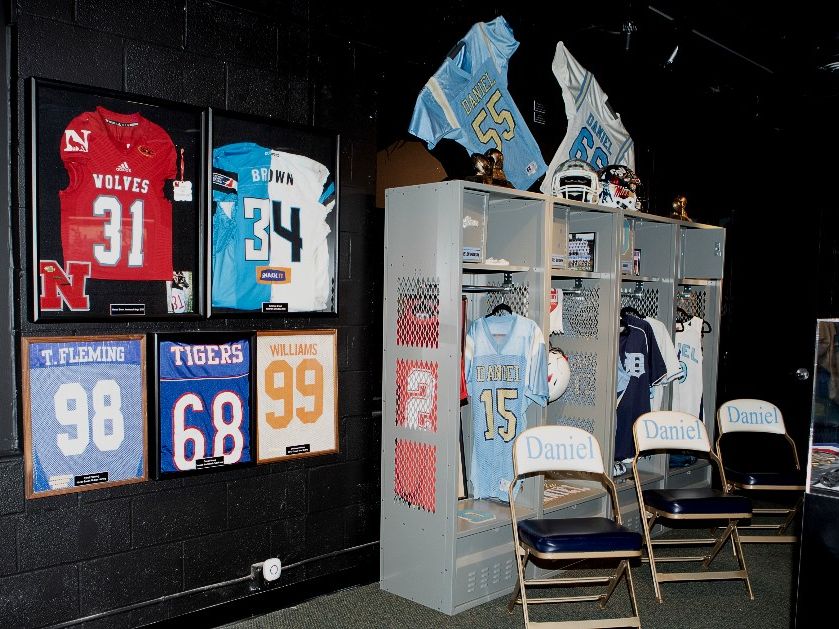 A sports memorabilia exhibit at the Clemson Area African American Museum displays framed football jerseys from local athletes mounted on dark walls alongside a recreated locker bay with Daniel High School uniforms, helmets, and folding chairs. The installation honors the athletic achievements of African American players from the Clemson area community