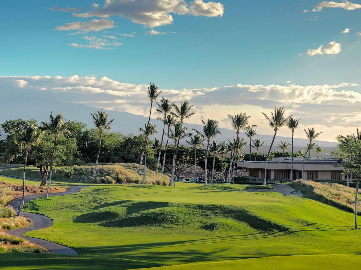 Tall swaying palm trees line the undulating fairways of Hapuna Golf Course on the Big Island of Hawaii, with the Mauna Kea volcano rising above the treeline in the background under a dramatic afternoon sky.