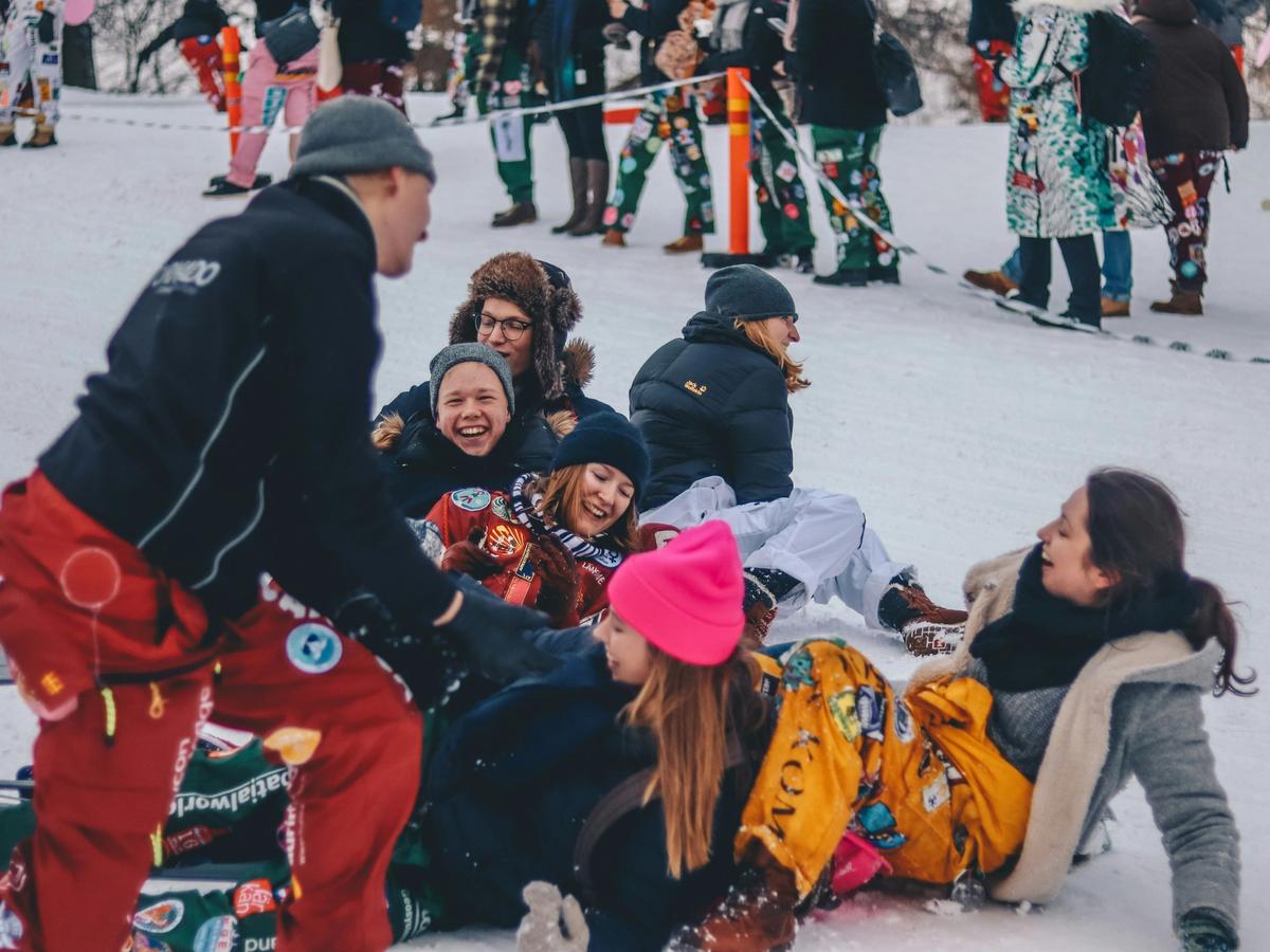 A joyful group of people dressed in colorful winter clothing laugh and play together in the snow, surrounded by others enjoying a festive outdoor winter event.