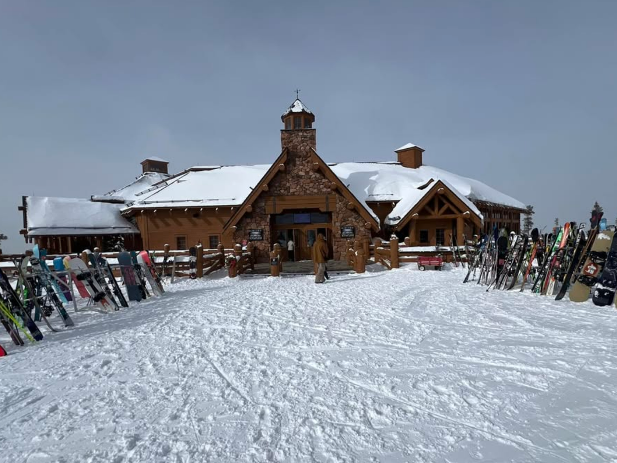 Rustic ski lodge with snow-covered roof surrounded by rows of skis and snowboards, set against a gray winter sky and snowy ground.