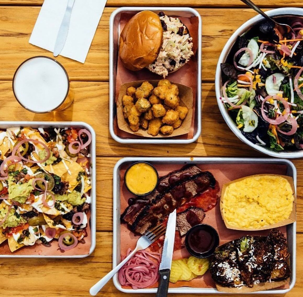 A table filled with barbecue favorites including smoked meats, nachos, salad, and classic Southern sides. The dishes are arranged on trays, perfect for sharing with friends or family. Highlands Smokehouse is known for its bold flavors and comforting Southern barbecue.