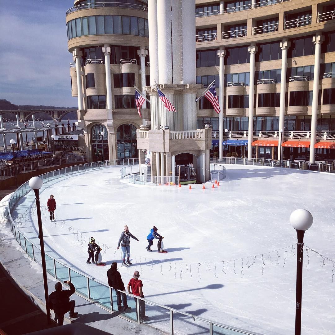 Families and children ice skate on the outdoor rink at Washington Harbour in Georgetown, DC, with American flags flying from the iconic waterfront building and the Key Bridge visible in the background on a crisp winter day.