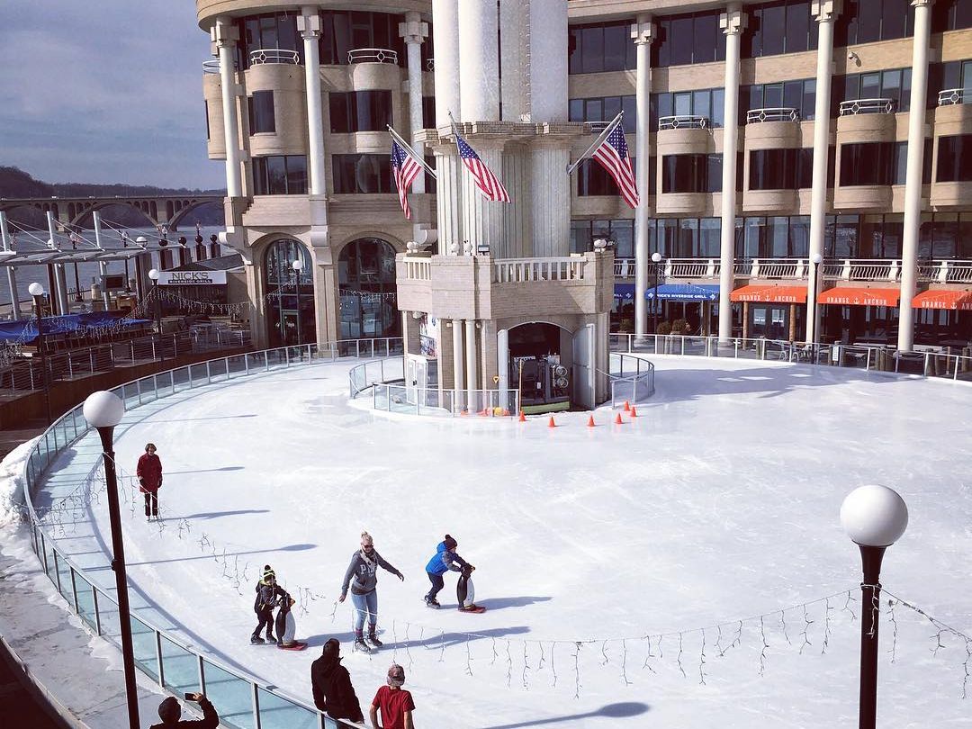 Families and children ice skate on the outdoor rink at Washington Harbour in Georgetown, DC, with American flags flying from the iconic waterfront building and the Key Bridge visible in the background on a crisp winter day.