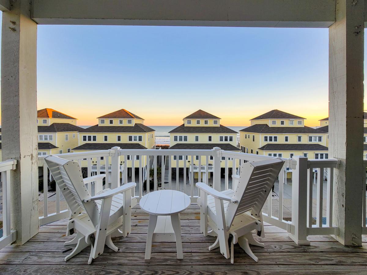 A serene balcony view at sunset featuring two white Adirondack chairs and a small table, overlooking rows of yellow beach houses with the ocean and colorful horizon in the distance.