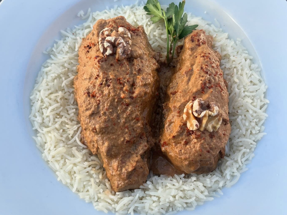 A plate of white rice topped with two pieces of chicken coated in a rich walnut and red pepper sauce, garnished with walnut halves and a sprig of parsley.