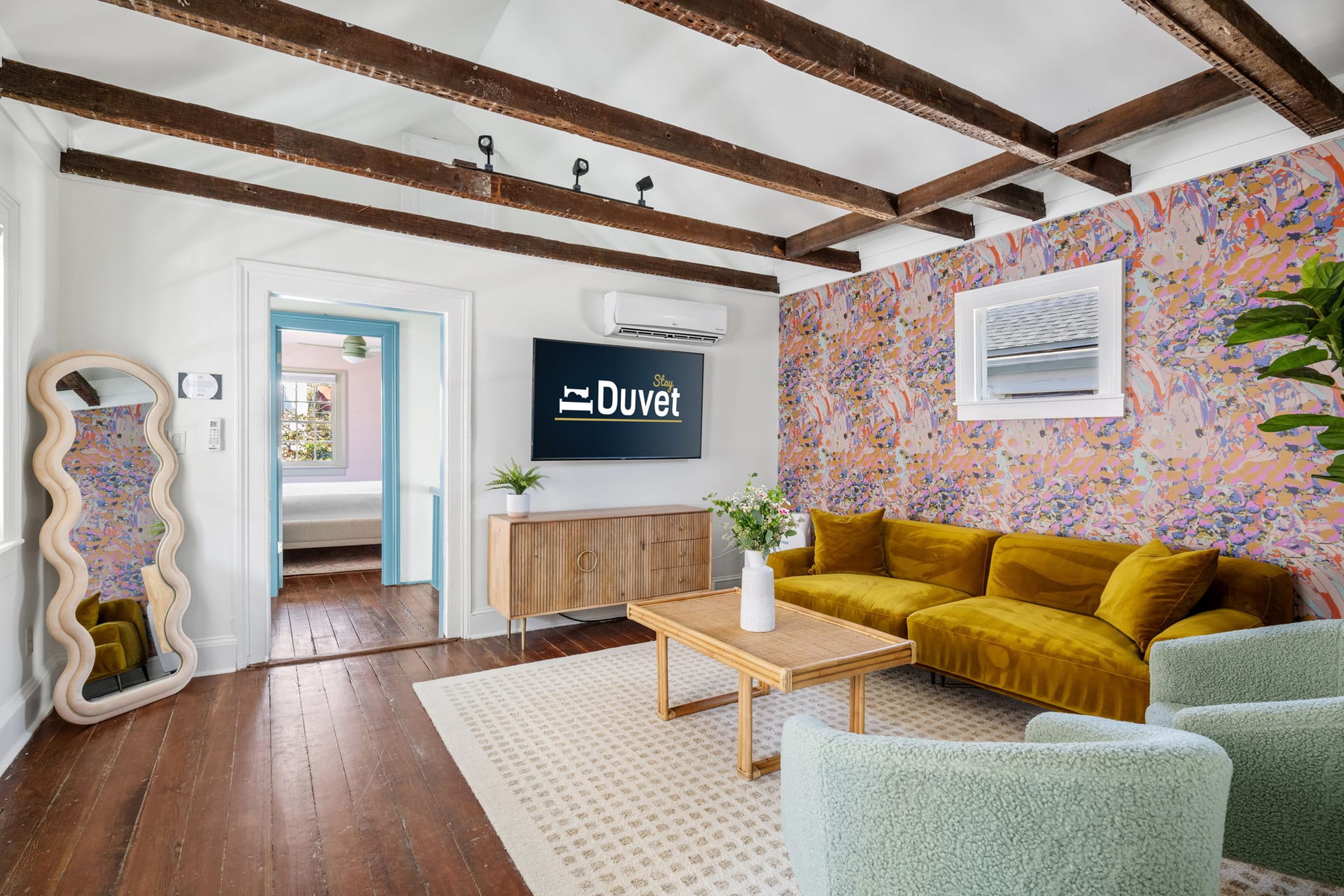 A bright and colorful living room in Charleston featuring exposed wood beams, a velvet mustard sofa, and playful patterned wallpaper. This cheerful home blends historic charm with modern design, perfect for a stylish downtown getaway.