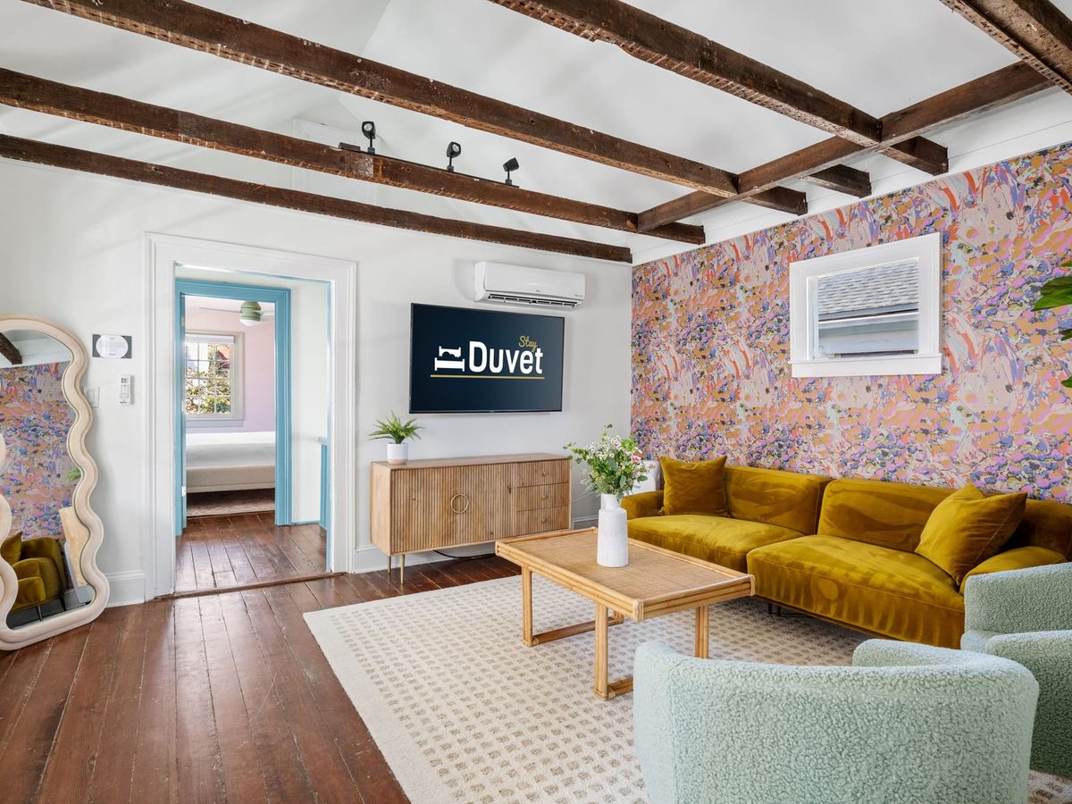 A bright and colorful living room in Charleston featuring exposed wood beams, a velvet mustard sofa, and playful patterned wallpaper. This cheerful home blends historic charm with modern design, perfect for a stylish downtown getaway.