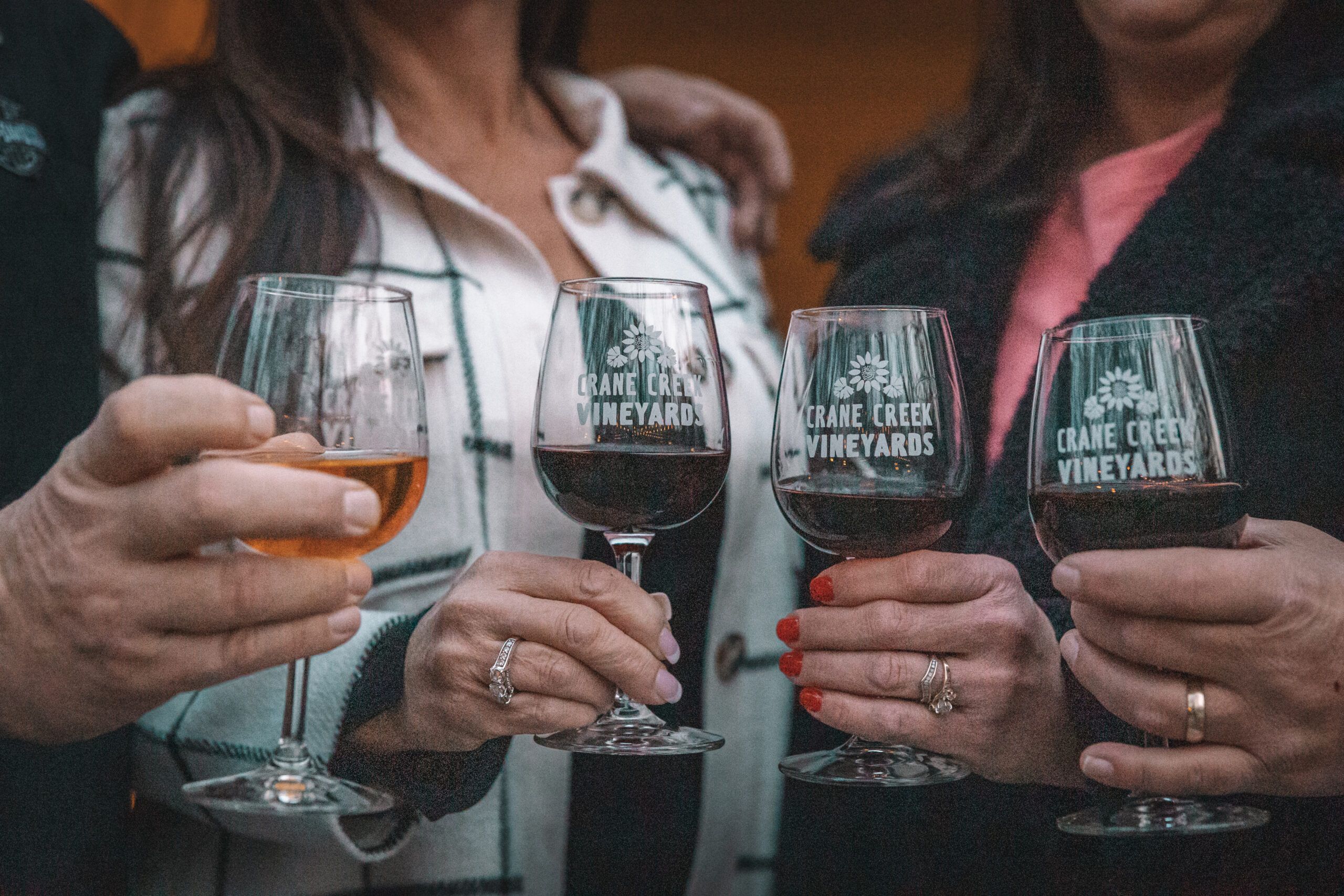 A group of friends holds glasses of wine from Crane Creek Vineyards, each glass filled with a different blend. Their hands and outfits add a warm, personal touch, showing a relaxed and fun tasting moment. It's a simple scene that feels friendly and inviting.