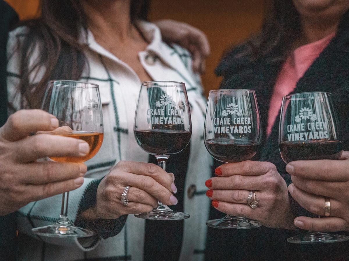 A group of friends holds glasses of wine from Crane Creek Vineyards, each glass filled with a different blend. Their hands and outfits add a warm, personal touch, showing a relaxed and fun tasting moment. It's a simple scene that feels friendly and inviting.