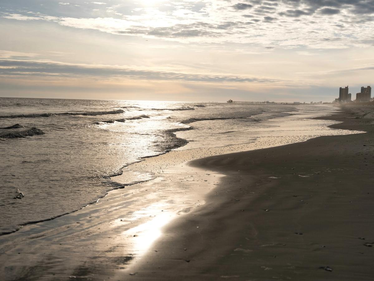 A quiet Myrtle Beach shoreline at low tide, with gentle waves reflecting soft morning light, a distant pier on the horizon, and high-rise buildings fading into the mist along the coast.