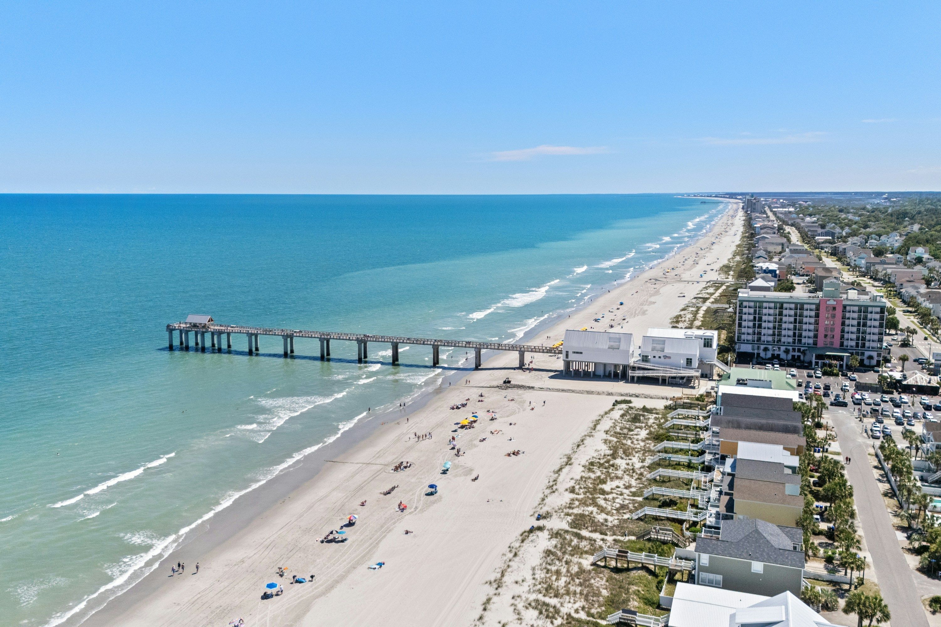 An aerial view of Myrtle Beach with a long pier extending into turquoise water, sandy shoreline dotted with beachgoers, and oceanfront homes and condos stretching along the coast under a clear blue sky.