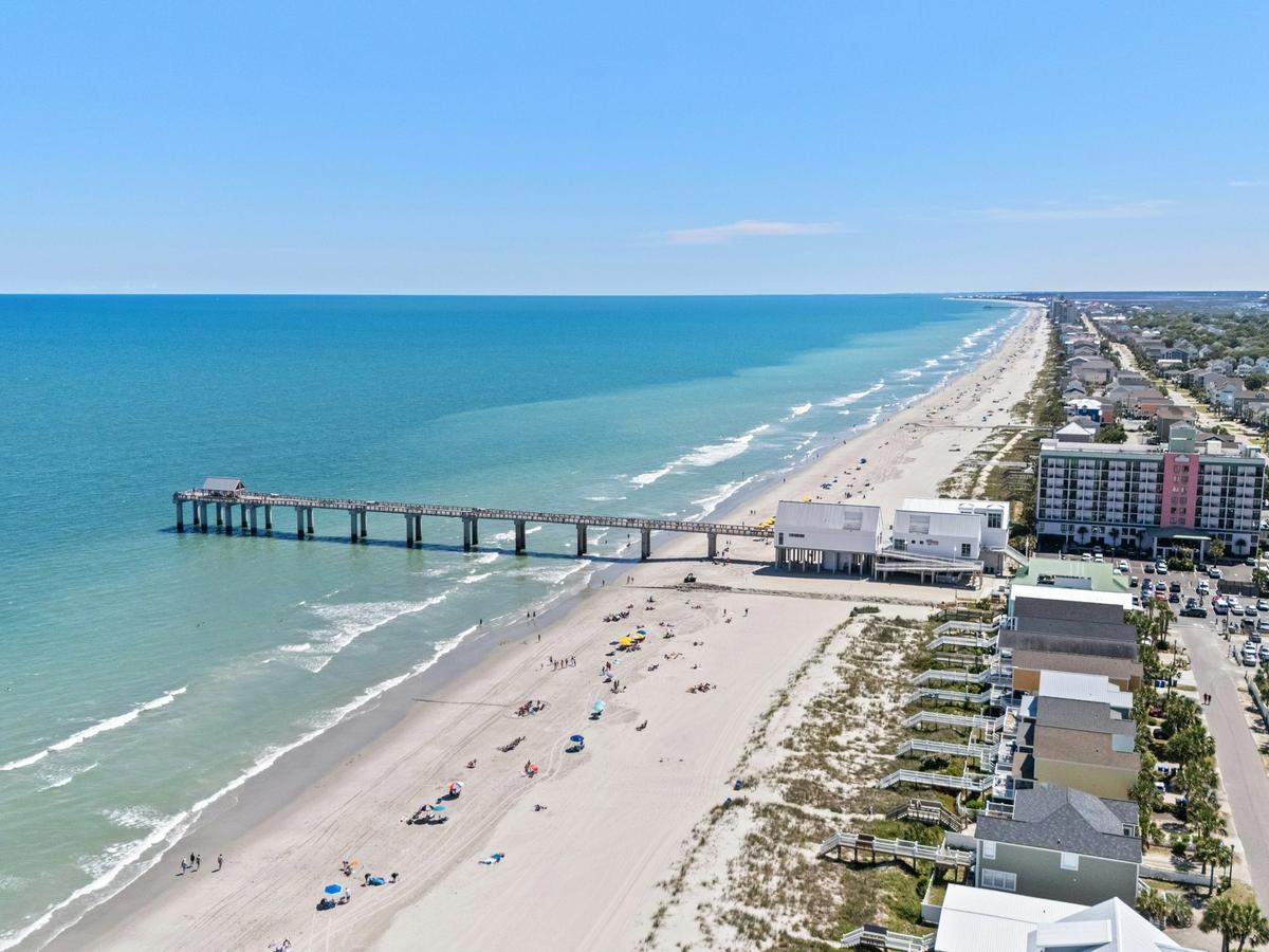 An aerial view of Myrtle Beach with a long pier extending into turquoise water, sandy shoreline dotted with beachgoers, and oceanfront homes and condos stretching along the coast under a clear blue sky.