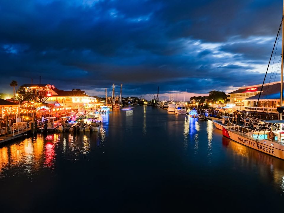 Shem Creek comes alive at night with glowing waterfront restaurants, lively bars, and boats docked along the water. The reflections of lights create a vibrant, welcoming atmosphere perfect for dining and nightlife.