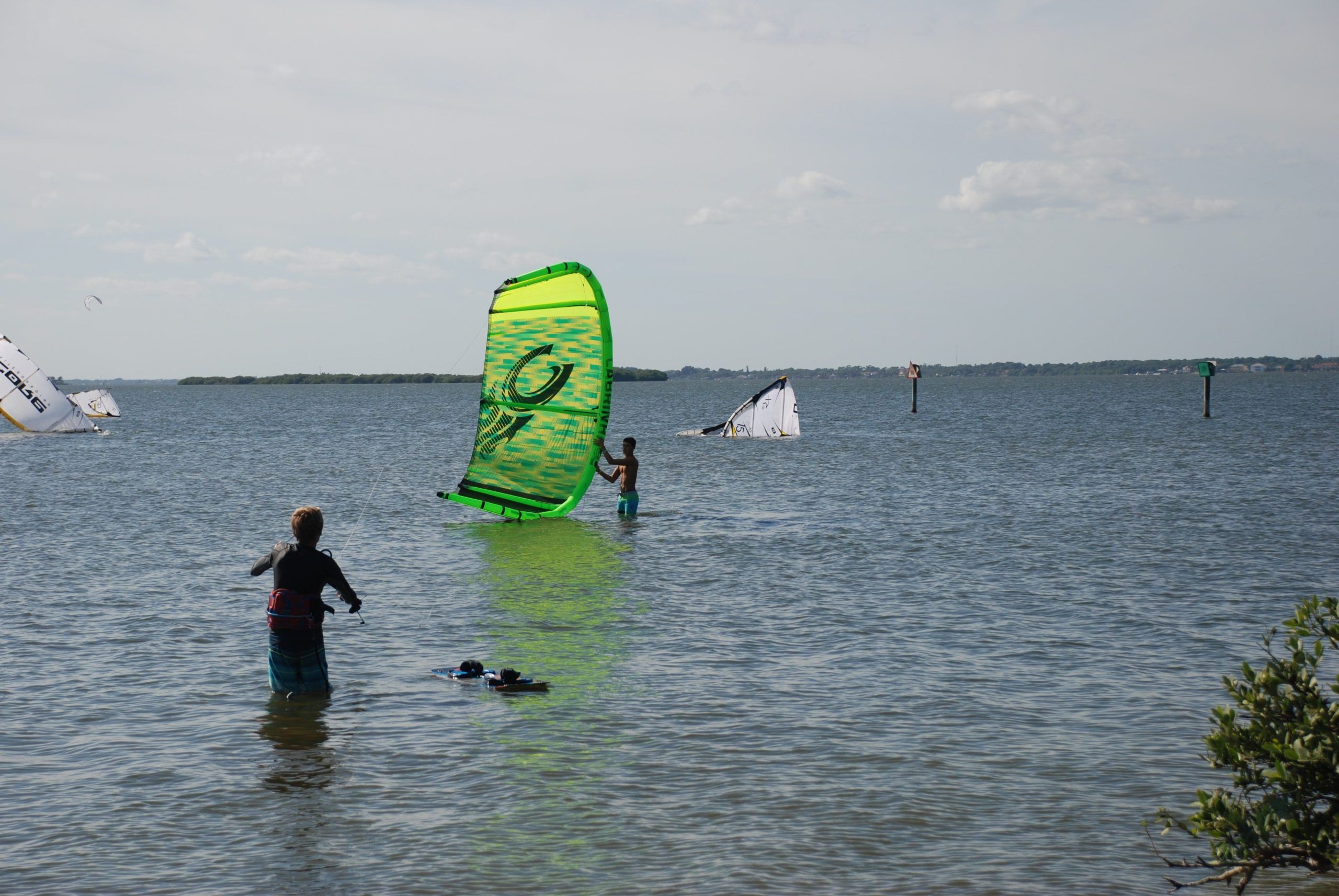 A kite surfer stands in shallow turquoise water, ready to launch a bright green sail into the wind. The calm sea and open sky make it the perfect setting for an adrenaline-filled day on the water.