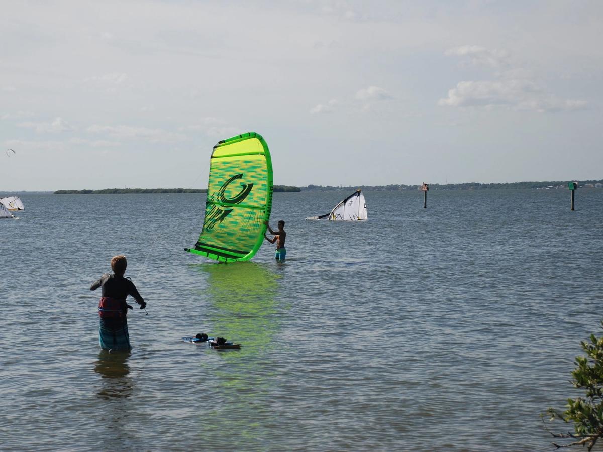 A kite surfer stands in shallow turquoise water, ready to launch a bright green sail into the wind. The calm sea and open sky make it the perfect setting for an adrenaline-filled day on the water.