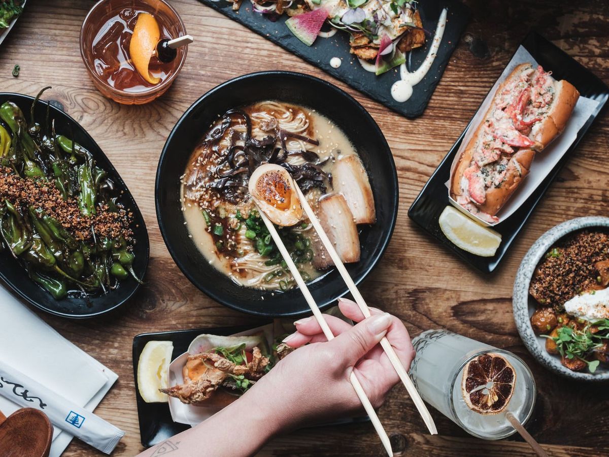 A steaming bowl of ramen at Domu, loaded with pork belly, egg, scallions, and mushrooms, surrounded by sides like shishito peppers and bao buns. Modern Japanese comfort food at its best