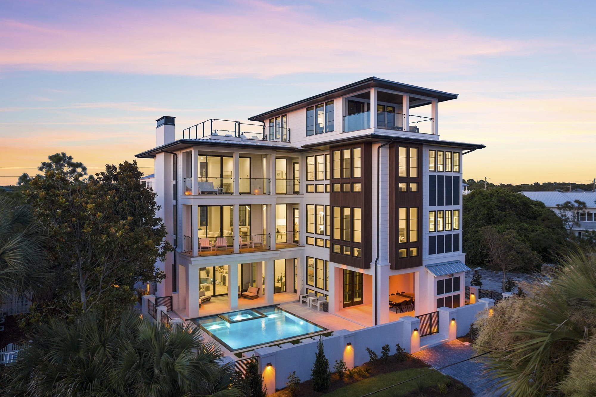 Above And Beyond on Florida’s 30A features several levels of covered balconies and large windows glowing at dusk. A lit pool and raised spa sit beside the home, framed by palm trees.