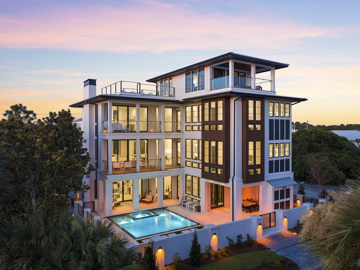Above And Beyond on Florida’s 30A features several levels of covered balconies and large windows glowing at dusk. A lit pool and raised spa sit beside the home, framed by palm trees.