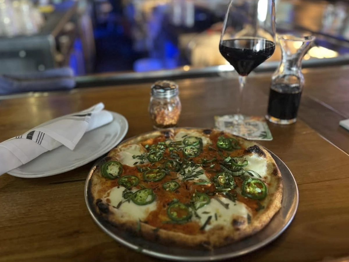 A wood-fired pizza topped with fresh mozzarella, sliced jalapeños, and basil sits on a bar counter next to a glass of red wine, a small carafe, and a shaker of red pepper flakes.