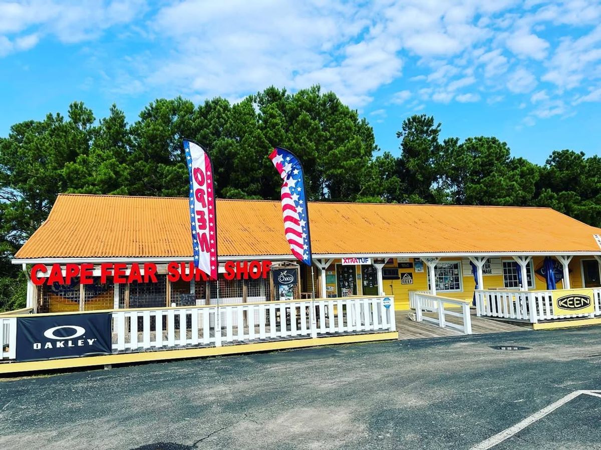 The bright yellow Cape Fear Surf Shop stands out with bold signs, waving flags, and gear for beach days. A fun, local favorite in Holden Beach.