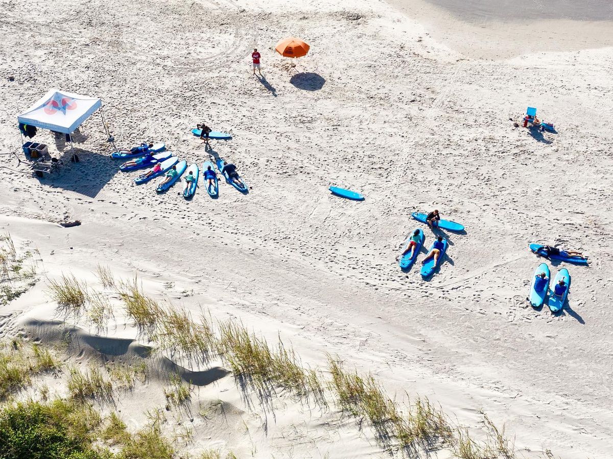 Aerial view of surf students lined up on the sand with blue surfboards during a group lesson at Folly Beach. Instructors guide beginners through the basics before heading into the water. Surf schools like this make learning fun and easy for all ages.