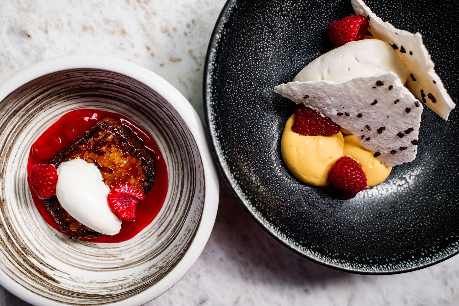 Two artfully plated desserts sit side by side, one a torched custard with whipped cream and raspberries over red coulis, the other a smooth mango cream with meringue shards and fresh berries in a speckled black bowl. The refined presentation highlights the kitchen's pastry craft.