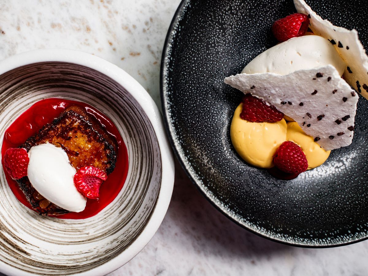 Two artfully plated desserts sit side by side, one a torched custard with whipped cream and raspberries over red coulis, the other a smooth mango cream with meringue shards and fresh berries in a speckled black bowl. The refined presentation highlights the kitchen's pastry craft.