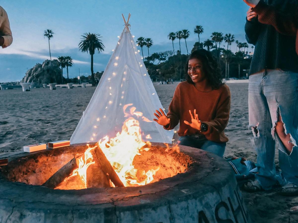 Sunset bonfire on the beach with friends, a glowing firepit, and a dreamy lighted teepee backdrop.
