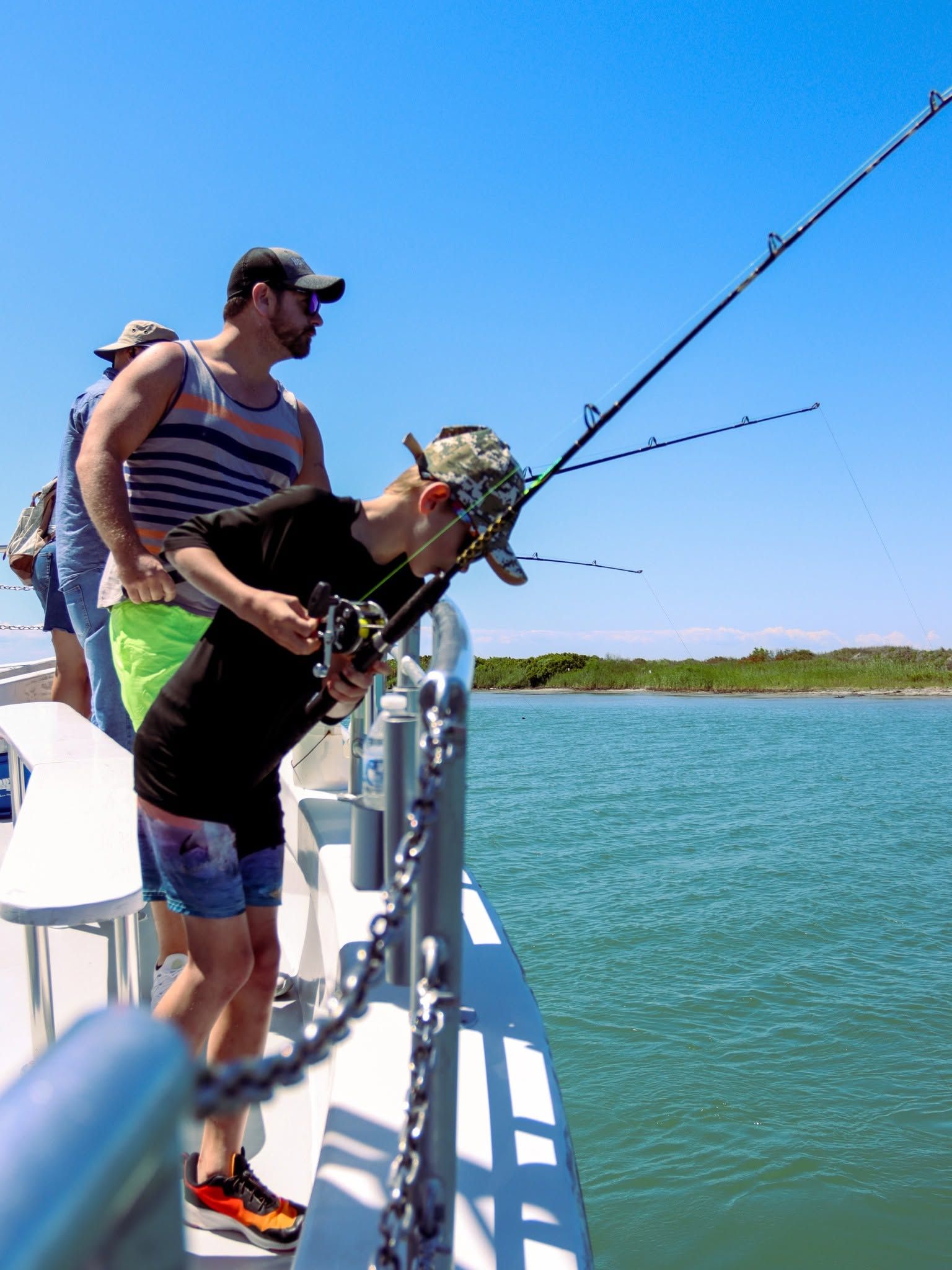 A young child in a camouflage cap leans over the railing of a fishing boat to reel in a line while an adult stands close behind for support. Calm turquoise water and a green marshy shoreline stretch out under a clear blue sky. 