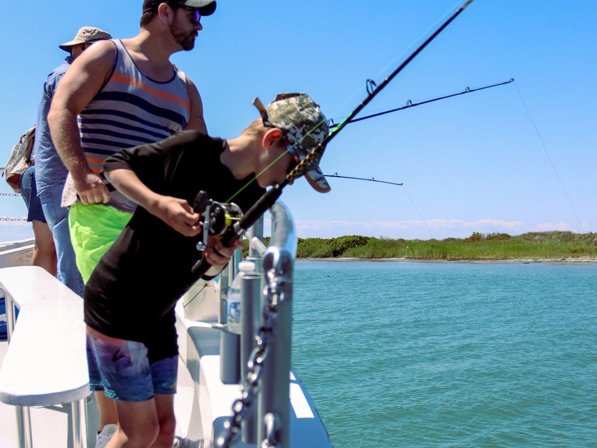A young child in a camouflage cap leans over the railing of a fishing boat to reel in a line while an adult stands close behind for support. Calm turquoise water and a green marshy shoreline stretch out under a clear blue sky.