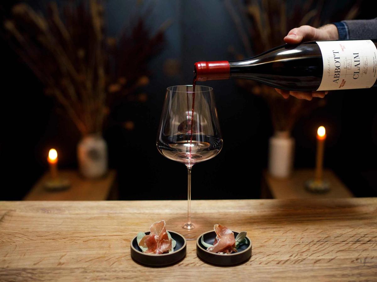 A bottle of Abbot Claim wine is carefully poured into a tall glass, set on a rustic wooden table with candlelight in the background. Small plates of cured meats add to the intimate tasting experience. The scene reflects the refined elegance of Oregon’s Willamette Valley wineries.