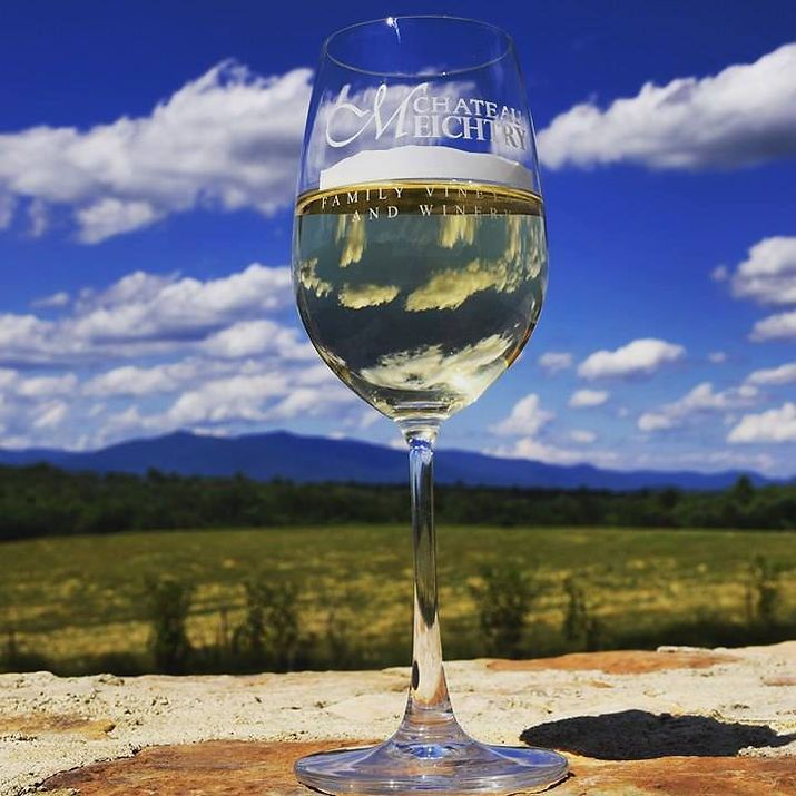 A glass of white wine from Chateau Meichtry stands against a backdrop of blue skies, open fields, and mountain views. The reflection in the glass adds a pretty touch to the scene. It captures the peaceful beauty of Georgia wine country.