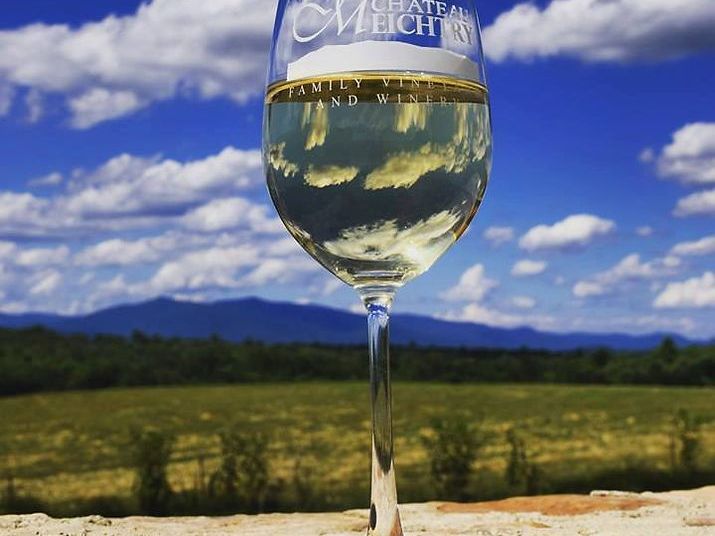 A glass of white wine from Chateau Meichtry stands against a backdrop of blue skies, open fields, and mountain views. The reflection in the glass adds a pretty touch to the scene. It captures the peaceful beauty of Georgia wine country.