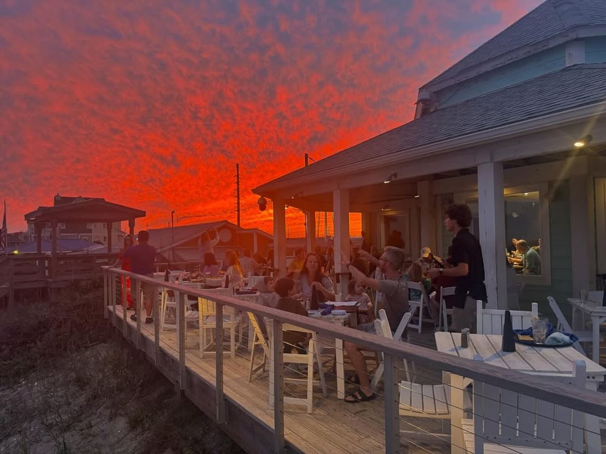 Families enjoy dinner on the deck of Daddy Mac’s Beach Grille under a blazing orange sunset. This oceanside restaurant is known for its seafood, good vibes, and unforgettable views.