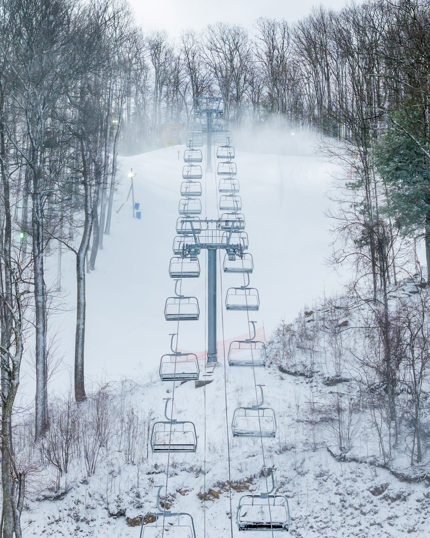 A snow-covered ski lift at Ober Gatlinburg stretches up a white powder-dusted slope flanked by bare winter trees and drifting snowmaker mist in the Great Smoky Mountains of Tennessee. The empty chairlift lines recede dramatically up the hill, capturing the serene stillness of a snowy mountain morning.