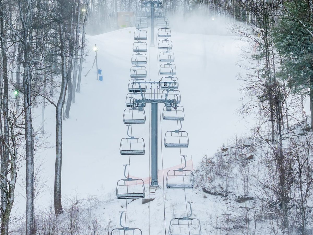 A snow-covered ski lift at Ober Gatlinburg stretches up a white powder-dusted slope flanked by bare winter trees and drifting snowmaker mist in the Great Smoky Mountains of Tennessee. The empty chairlift lines recede dramatically up the hill, capturing the serene stillness of a snowy mountain morning.