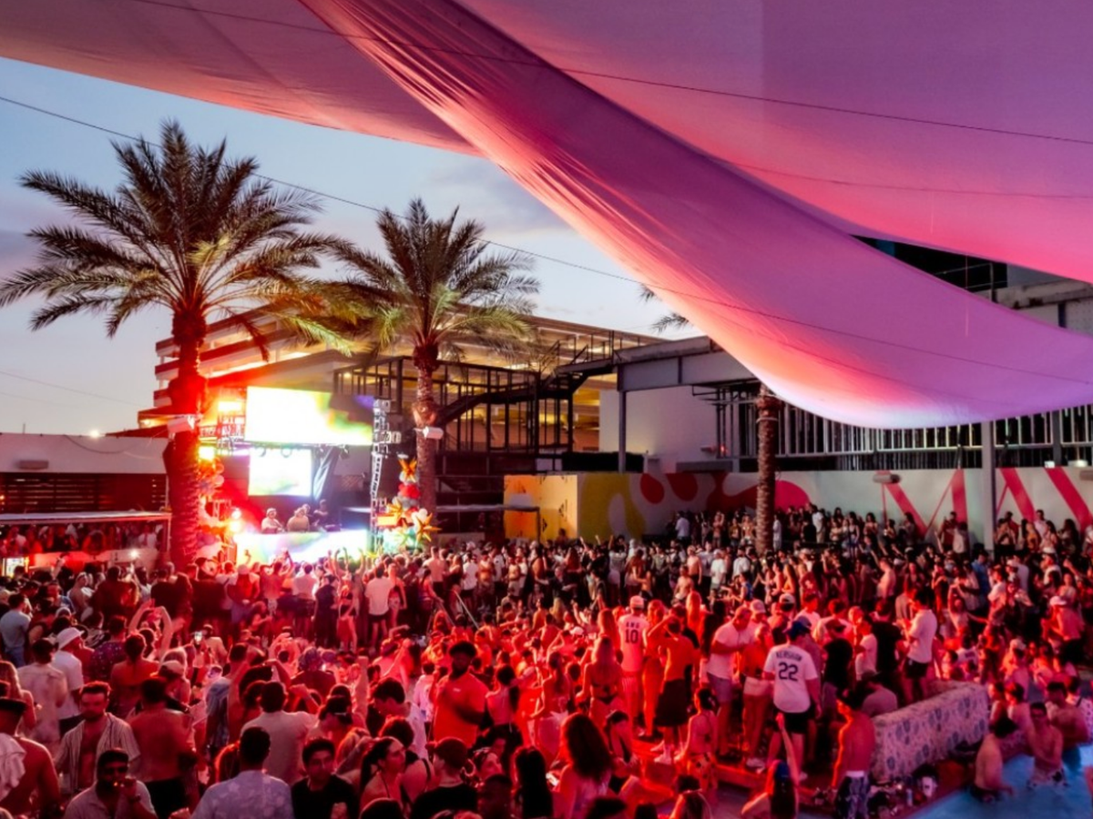 A lively crowd enjoys an outdoor sunset pool party with a DJ, colorful lighting, and palm trees under a large fabric canopy.